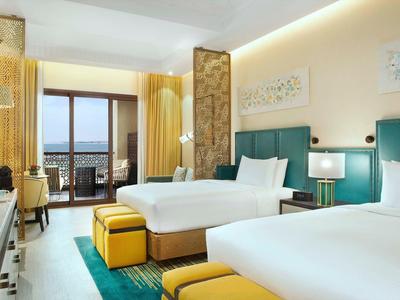 Helles Hotelzimmer mit zwei Doppelbetten, türkisfarbenen Kopfstützen, gelben Hockern und Balkonblick.