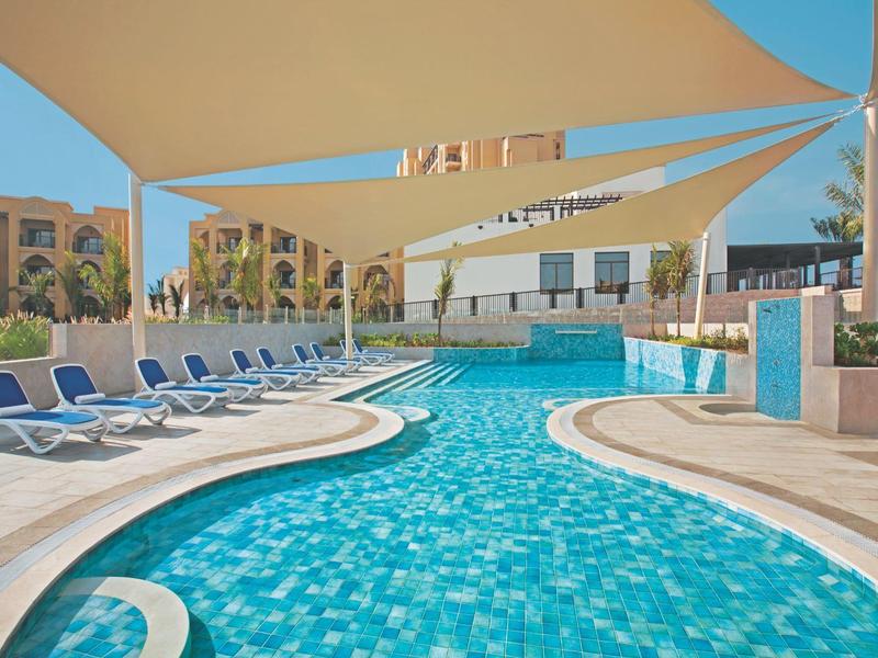 Freizeitpool mit blauen Liegestühlen und großen Sonnensegeln über beigem Boden.