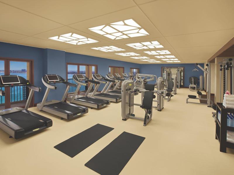 Großer Fitnessraum mit Laufbändern, Kraftgeräten und matten, beleuchtet durch große Deckenfenster.