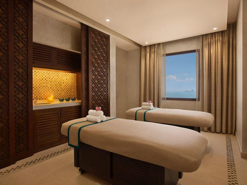 Zimmer mit zwei Massageliegen, warmem Licht und braunen, gemusterten Holzelementen an der Wand.