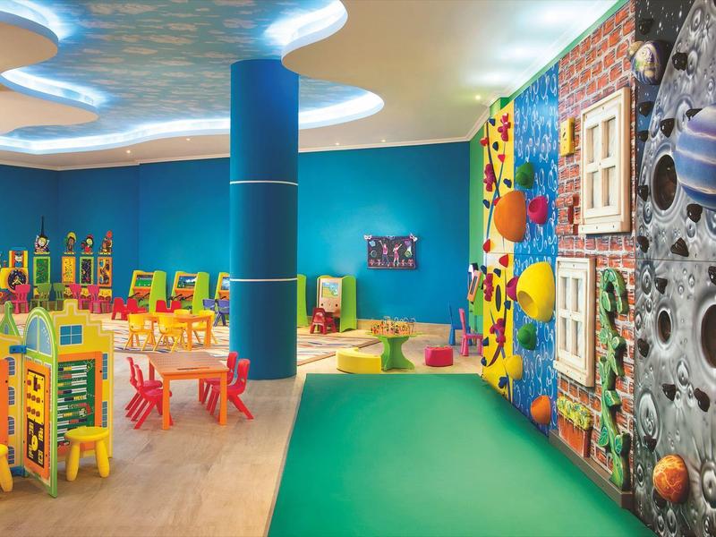 Bunter Kinderspielraum mit Kletterwand, Tischen, Stühlen und hellen, lebhaften Farben.