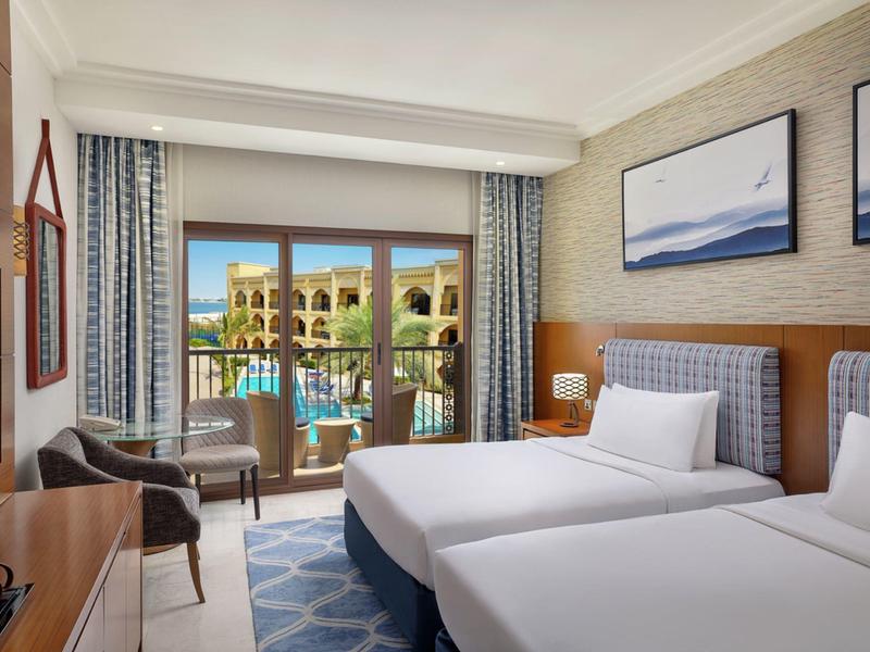 Hotelzimmer mit zwei Einzelbetten, Balkon und Blick auf einen Pool und Gebäude.