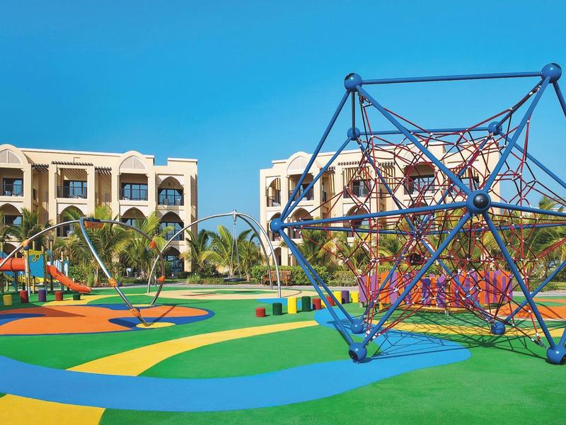 Bunt gestalteter Spielplatz mit Klettergerüst und Wohngebäuden im Hintergrund bei klarem Himmel.