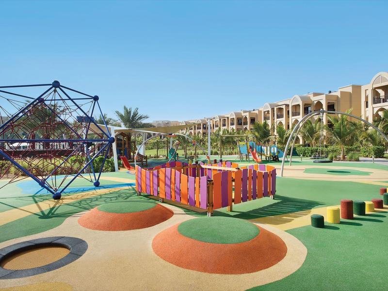 Bunter Spielplatz mit Klettergerüst, Hügeln, Balancierstangen und Rutschen vor Wohngebäuden.