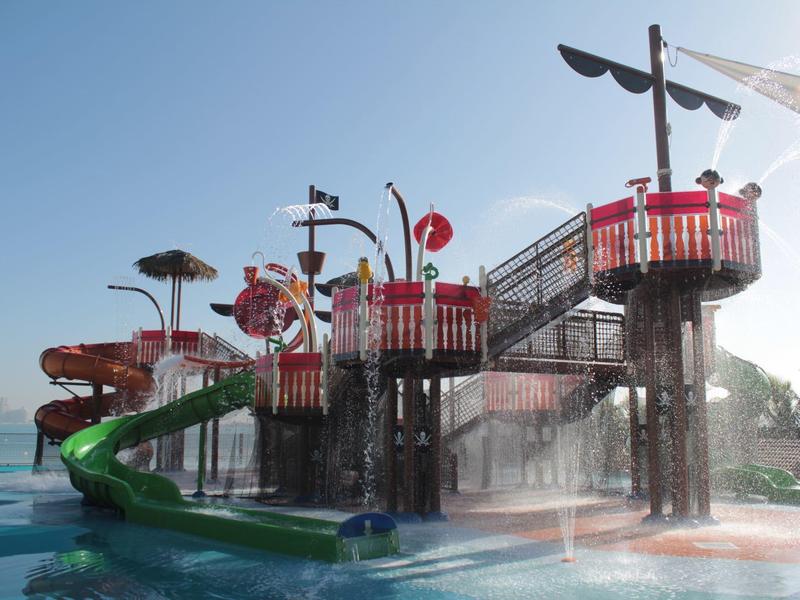 Wasserpark mit großen Rutschen, roten Geländern und Sprühdüsen bei klarem Himmel.