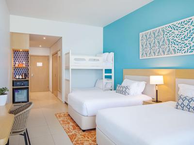 Modern eingerichtetes Hotelzimmer mit Doppelbett, Etagenbett, Schreibtisch und leuchtend blauer Wand.