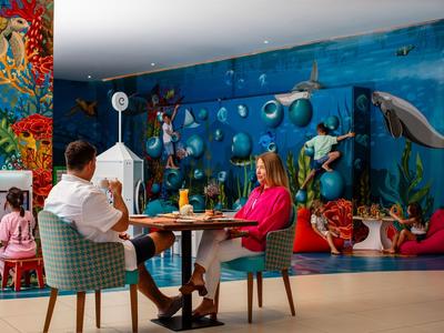 Dos adultos sentados en una mesa frente a un mural submarino colorido con peces y coral.