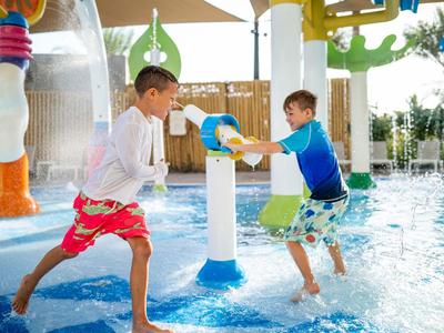 Dos niños juegan con rociadores de agua en un parque acuático bajo sombrillas.