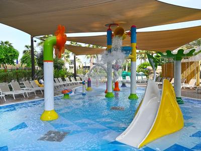 Zona de juegos para niños con toboganes y juegos de agua coloridos bajo toldos junto a la piscina del hotel.