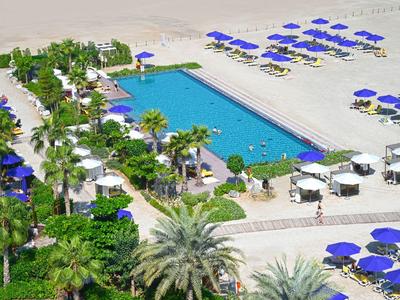 Gran área de piscina con sombrillas azules, arena blanca y vegetación tropical junto al hotel.