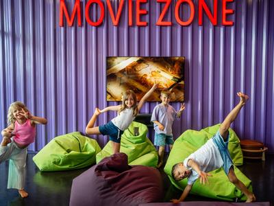 Niños bailan y juegan sobre pufs coloridos en una habitación con paredes moradas y cartel "MOVIE ZONE".