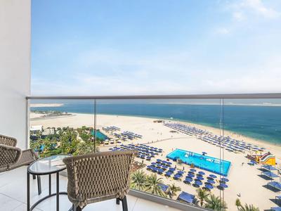 Balcón con sillas y mesa con vista a la playa, piscina y mar bajo un cielo azul.
