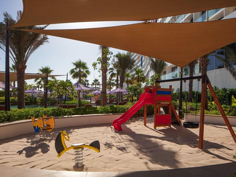 Overdekte kinderspeelplaats met glijbaan en wipkippen voor een hotel en palmbomen.