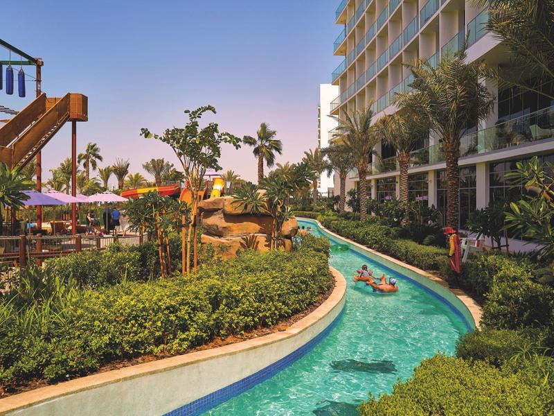 Buitenzwembadgebied met slow river, tropische planten en hotelgebouw op de achtergrond bij zonsondergang.