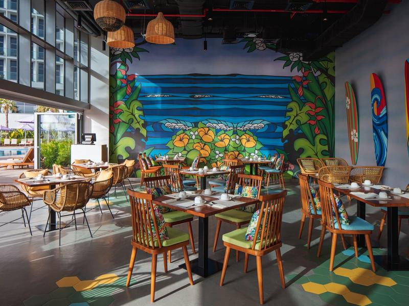 Modern restaurant met kleurrijke houten stoelen, muurschildering en grote ramen met uitzicht naar buiten.