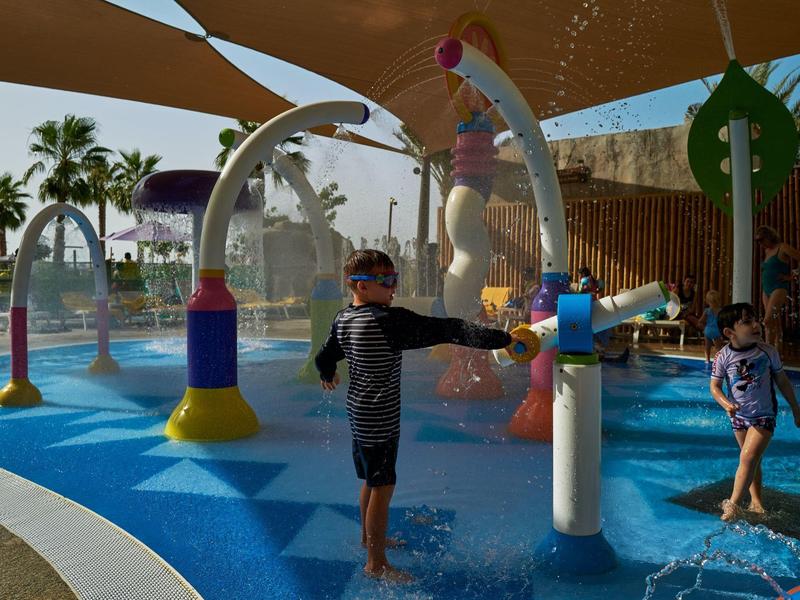 Kinderen spelen in een kleurrijk buitenspeelwatergebied met palmbomen op de achtergrond.