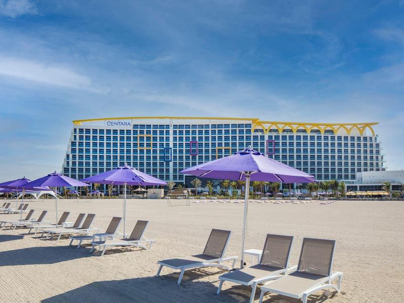 Meerlagig hotel op de achtergrond met strandstoelen en paarse parasols op het zandstrand.