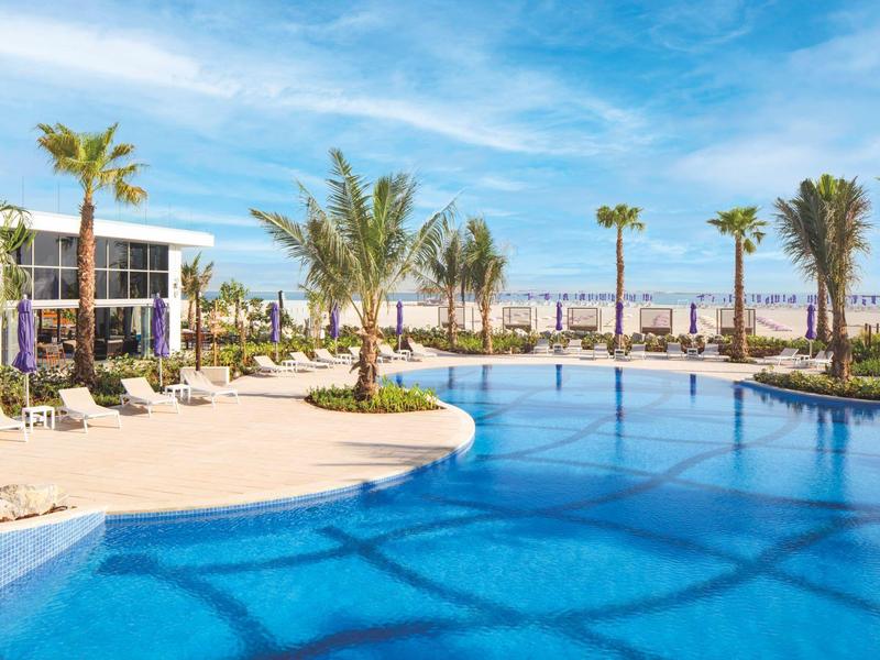 Groot helder zwembad met palmbomen en ligstoelen onder een blauwe lucht in een resort.