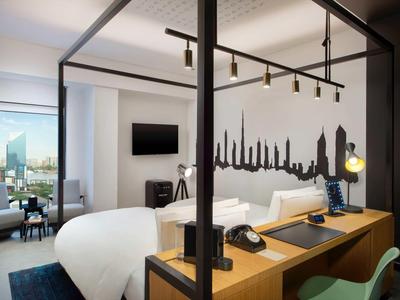 Modern hotel room with double bed, city skyline wall decor, and workspace with computer.