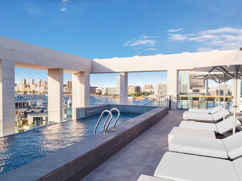 Rooftop pool with city views, white loungers, and umbrellas under a blue sky.