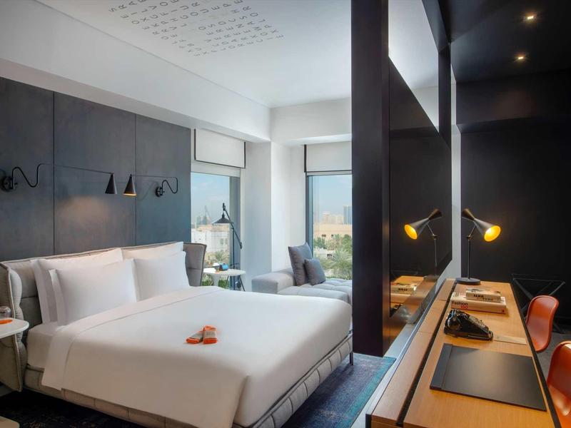 Modern hotel room with double bed, desk, and seating area in neutral tones.