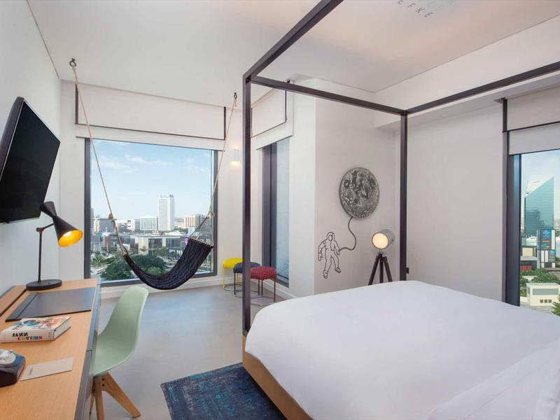 Modern hotel room with double bed, desk, TV, and large windows with city views.