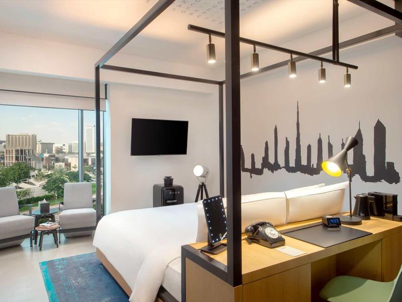 Modern hotel room with canopy bed, desk, and city view through large windows.