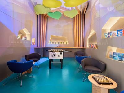 Moderner Spielraum mit bunten Luftballons, blauem Boden, blauen Sesseln und einem Kicker in der Mitte.