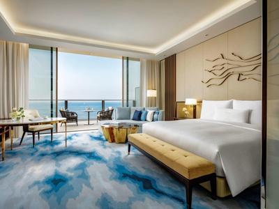 Helles Hotelzimmer mit großem Bett, blauem Teppich und Meerblick durch große Fenster.