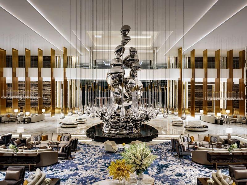 Großer, heller Hotel-Lobbybereich mit moderner Skulptur, Sitzgruppen und blauen Teppichen.