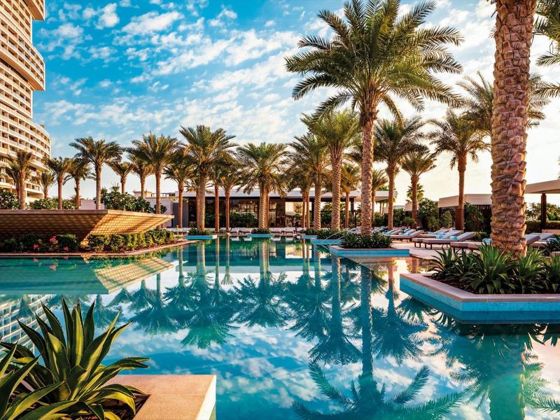 Schwimmbad mit klar blauem Wasser, umgeben von Palmen und bunten Blumen unter blauem Himmel.