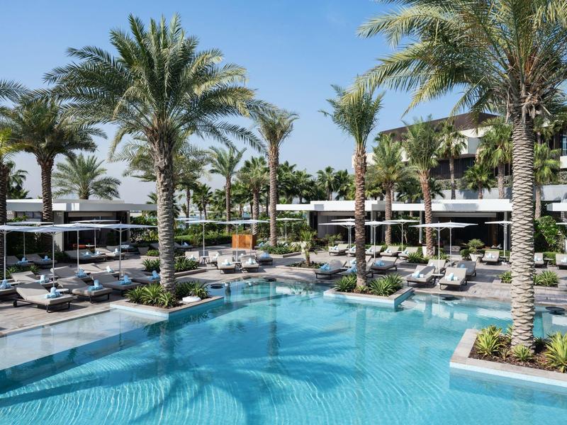 Moderner Poolbereich mit klar blauem Wasser, Palmen und Liegestühlen unter klarem Himmel.