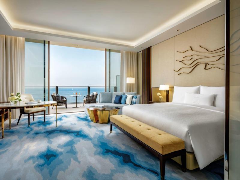 Helles Hotelzimmer mit großem Bett, blauem Teppich und Meerblick durch große Fenster.