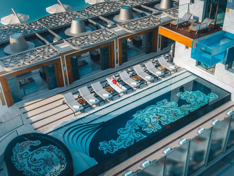 Moderner Außenpool mit aufwändigen blauen Fliesenmustern und Liegen auf weißem Holzdeck.