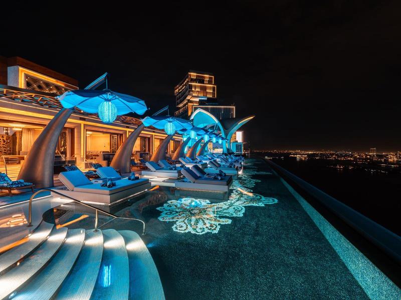 Luxuriöser Poolbereich mit blauer Beleuchtung, Liegestühlen und Stadtlichtern bei Nacht.