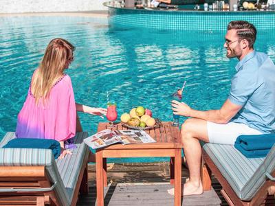 A couple relaxes by a pool on lounge chairs with drinks on a table between them.