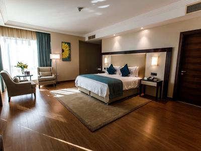 Bright hotel room with double bed, seating area, wooden floor, and natural light from windows.