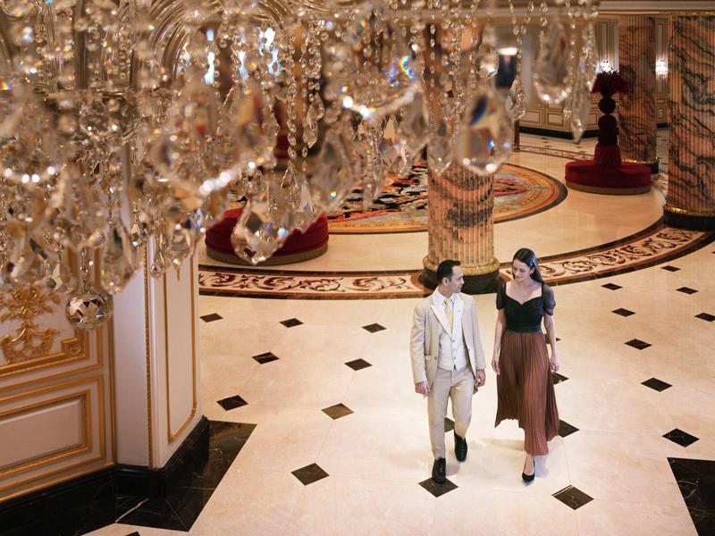 Zwei Personen gehen in luxuriösem Saal mit glänzendem Marmorboden und großem Kristallleuchter.