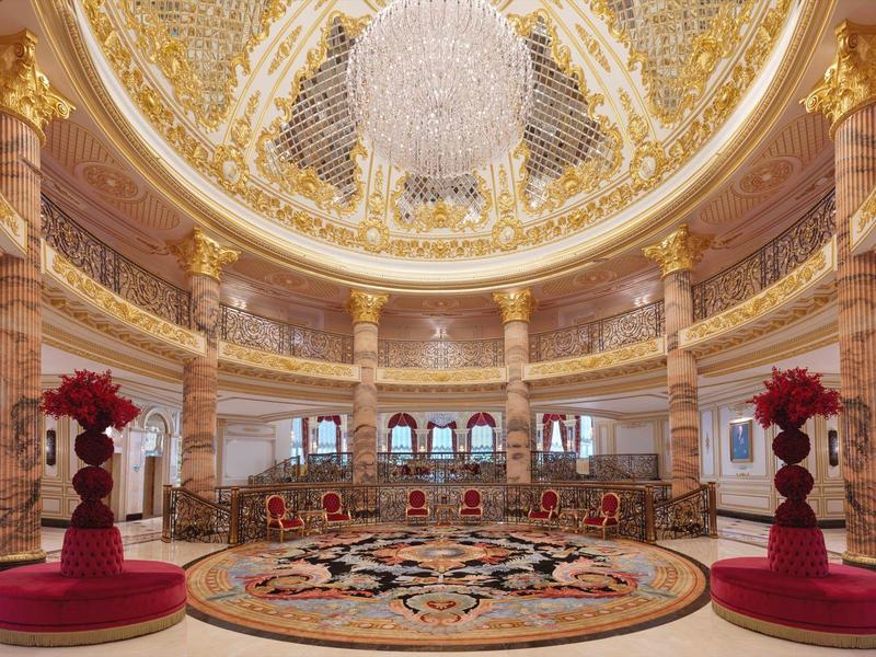 Prächtiger Saal mit goldverzierten Säulen, großem Kronleuchter und farbenreichem Teppich.