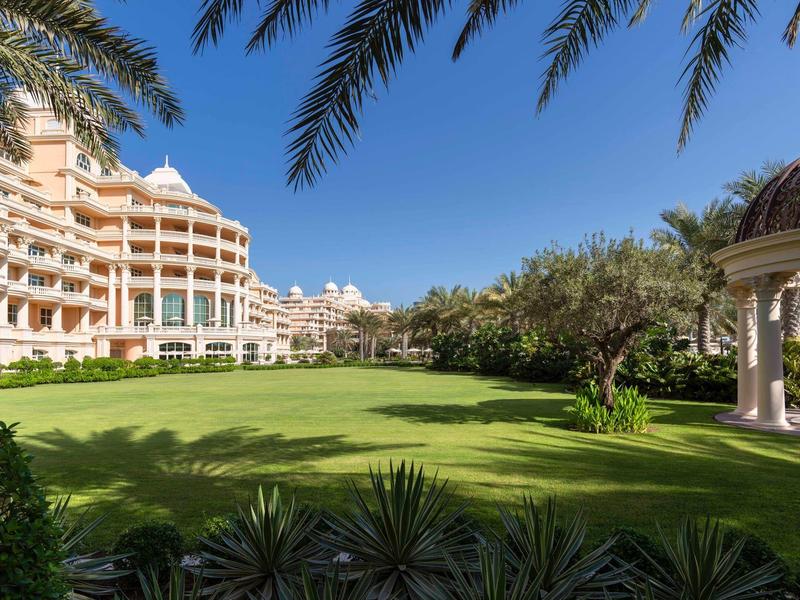 Großer, grüner Garten mit Palmen, Gebäuden links und Pergola rechts unter blauem Himmel.