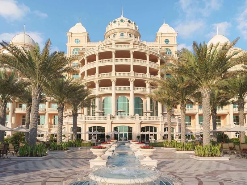 Großes Gebäude mit Kuppeln, Palmen und Springbrunnen im Vordergrund unter blauem Himmel.