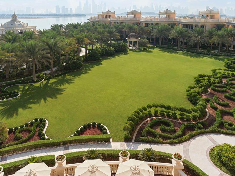 Große, gepflegte grüne Rasenfläche mit Palmen und geschwungenen Blumenbeeten in einer Gartenanlage.