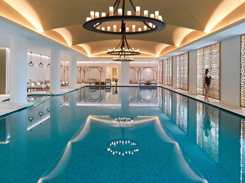 Elegantes Hallenbad mit blauem Wasser, warmem Licht, runden Kronleuchtern und modernen Wandverkleidungen.