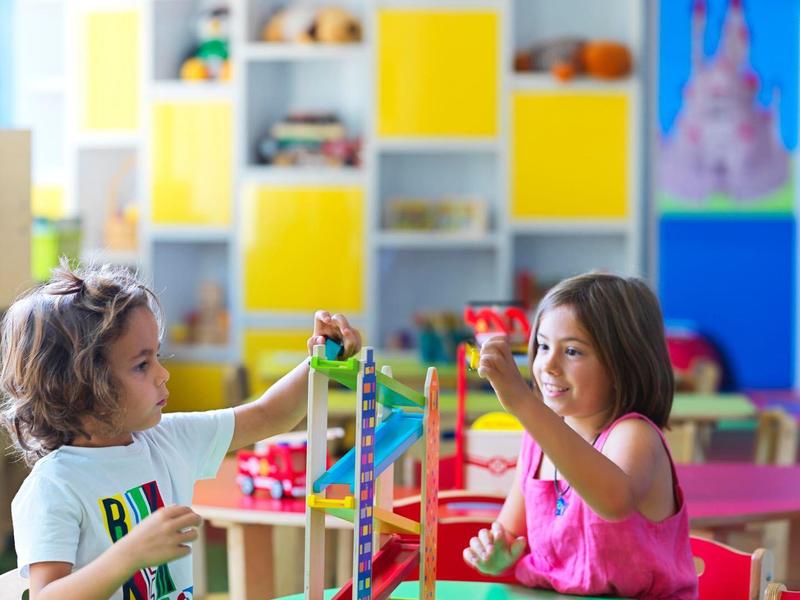 Zwei Kinder spielen an einem bunten Tisch in einem Zimmer mit gelben Regalen und Spielzeug.