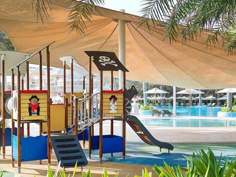 Bunter Kinderspielplatz mit Rutsche unter Sonnensegel neben großem Außenpool und Palmen.