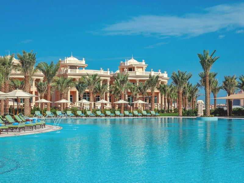 Großer Pool vor einem Hotel mit Palmen, Liegen und Sonnenschirmen unter blauem Himmel.