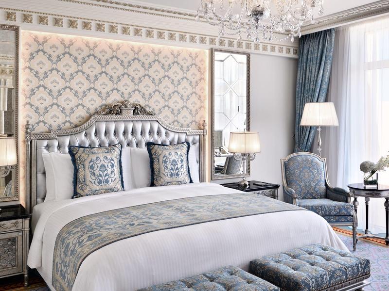 Elegantes Schlafzimmer mit großem Bett, gemusterten Kissen, Barocktapete und Sitzbereich am Fenster.