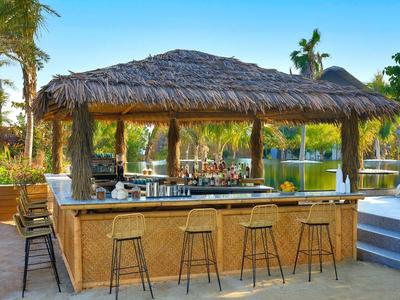 Bar al aire libre con techo de paja, taburetes junto a una piscina y palmeras.