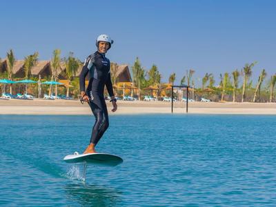 Person surfing on a foil board in clear blue water near a hotel beach with umbrellas.