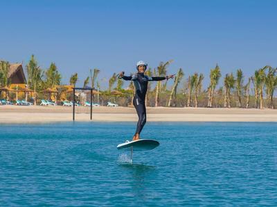 Person in wetsuit riding a hydrofoil surfboard over blue water on a sunny beach.